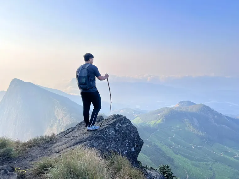 Joalex standing on a rocky summit above tea plantations and cloud-wrapped hills, leaning on a trekking stick